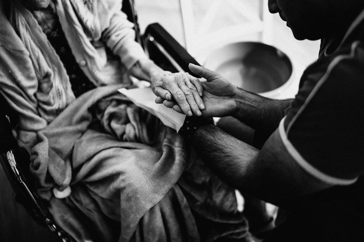 A caregiver holds the hand of an older woman seated in a wheelchair