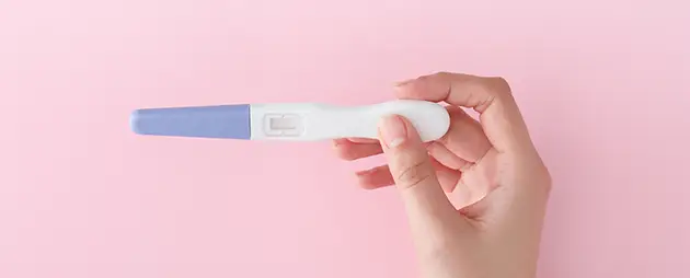 A hand holding a pregnancy test against a pink background